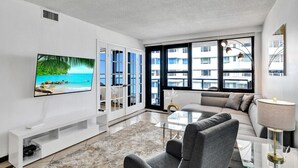 A modern living room with a flat-screen TV, a sofa, a glass coffee table, and a large window with a view of buildings.