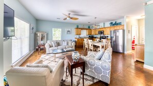 A spacious living room with a sofa, coffee table, and a TV. A kitchen with wooden cabinets and a dining area is visible in the background.