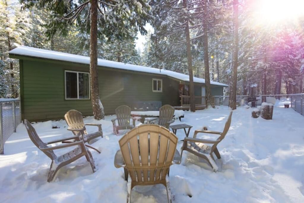 Una baita con tetto innevato, circondata da alberi. In primo piano si vedono diverse sedie di legno e un tavolino.