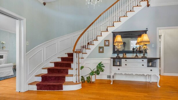 A grand staircase with a wooden handrail and red carpeted steps, leading to a landing with a white console table and a mirror. - Beland Mansion Getaway – Spacious Colonial with Game Room & Lounge (Fort Smith)