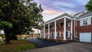 A two-story brick building with a covered front porch supported by columns, a garage, and a large tree in the foreground. - Beland Mansion Getaway – Spacious Colonial with Game Room & Lounge (Fort Smith)