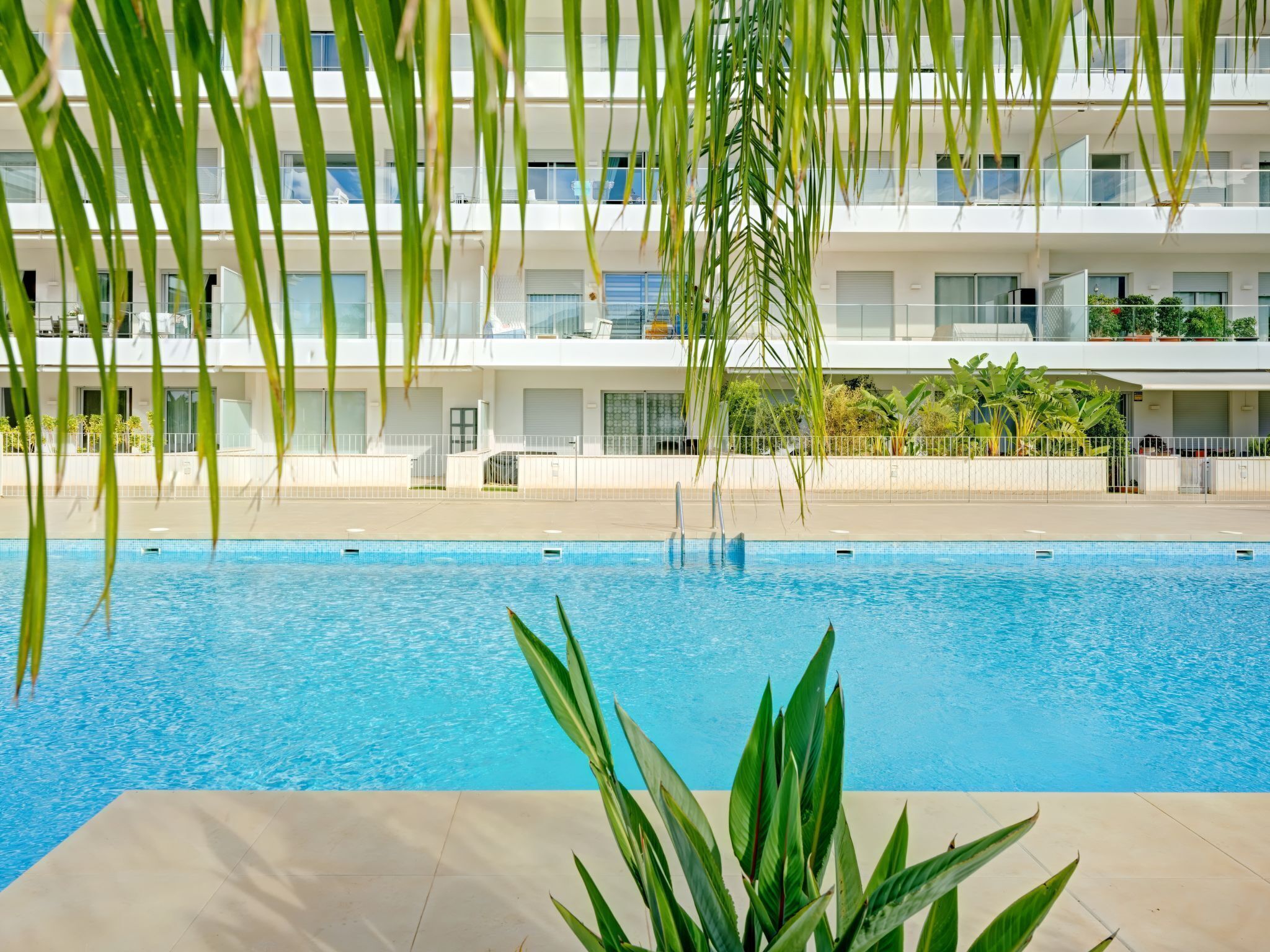 A swimming pool with clear blue water, surrounded by a tiled deck. In the background, there is a multi-story building with balconies and potted plants.