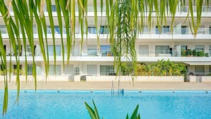 Ein Swimmingpool mit klarem, blauem Wasser, umgeben von einer gefliesten Liegefläche. Im Hintergrund ein mehrstöckiges Gebäude mit Balkonen und Blumentöpfen.