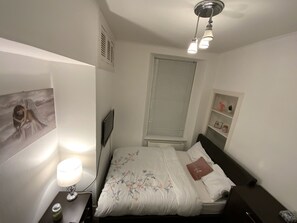 A bedroom with a bed, bedside table, lamp, wall art, window, and built-in shelving.