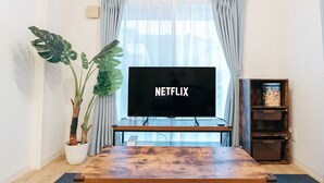 En stue med et fladskærms-tv, der viser "NETFLIX", et træ sofabord, en potte med en monstera plante og en reol med en børnebog.