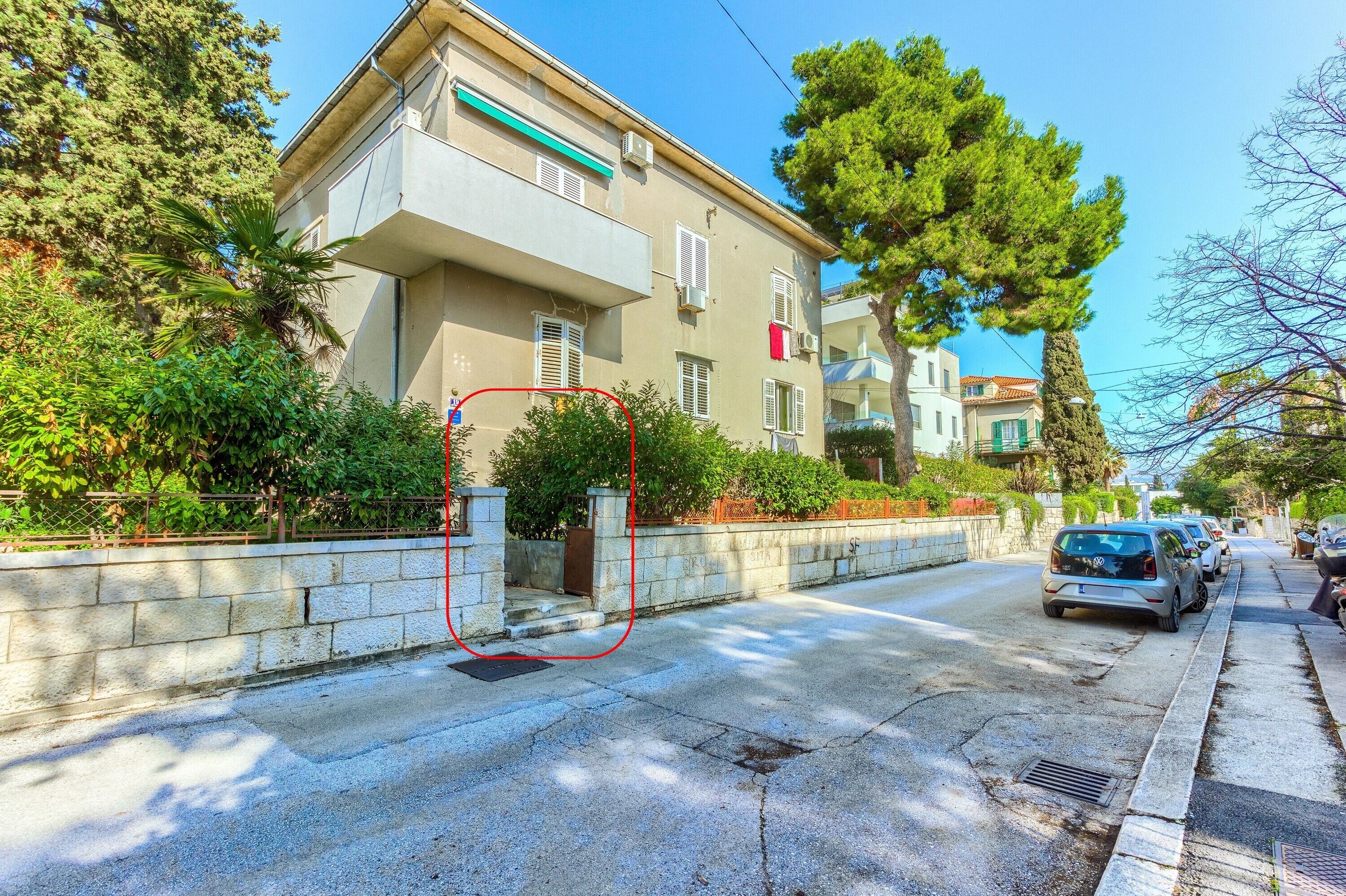 A residential street with a two-story building, a gated entrance, parked cars, and surrounding greenery.