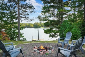 Un espace extérieur aménagé au bord du lac, avec un foyer, entouré de chaises Adirondack, offrant une vue sur un lac paisible et une forêt dense.