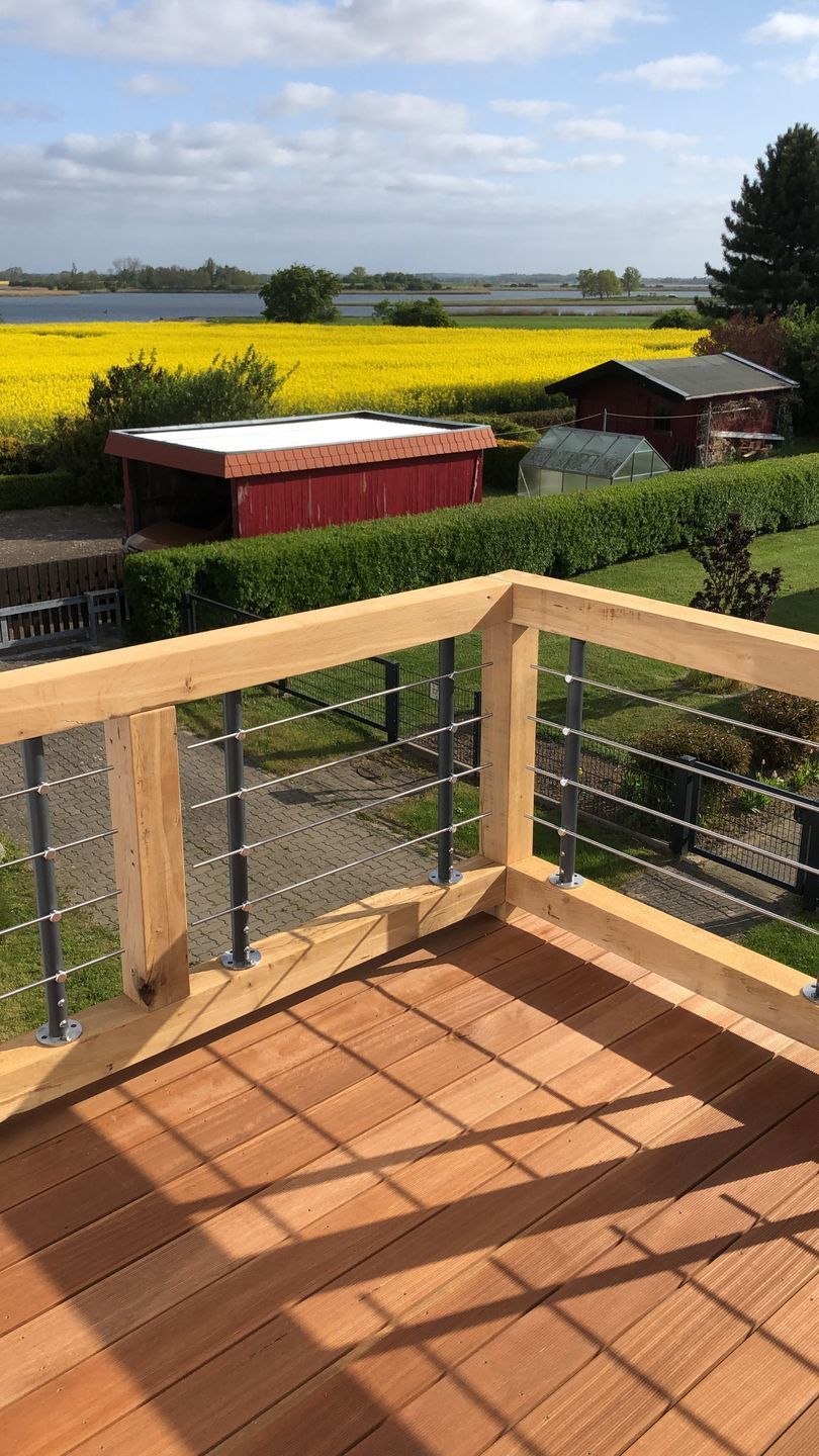 En träveranda med ett metallräcke som vetter ut mot ett fält med gula blommor och en röd lada.