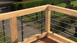 Eine Holzterrasse mit Metallgeländer, die auf ein Feld mit gelben Blumen und ein rotes Bauernhaus blickt.