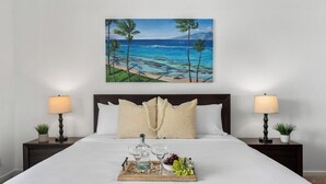 Habitación de hotel con cama, dos mesitas de noche, una bandeja con bebidas y aperitivos, y un cuadro de una playa en la pared.