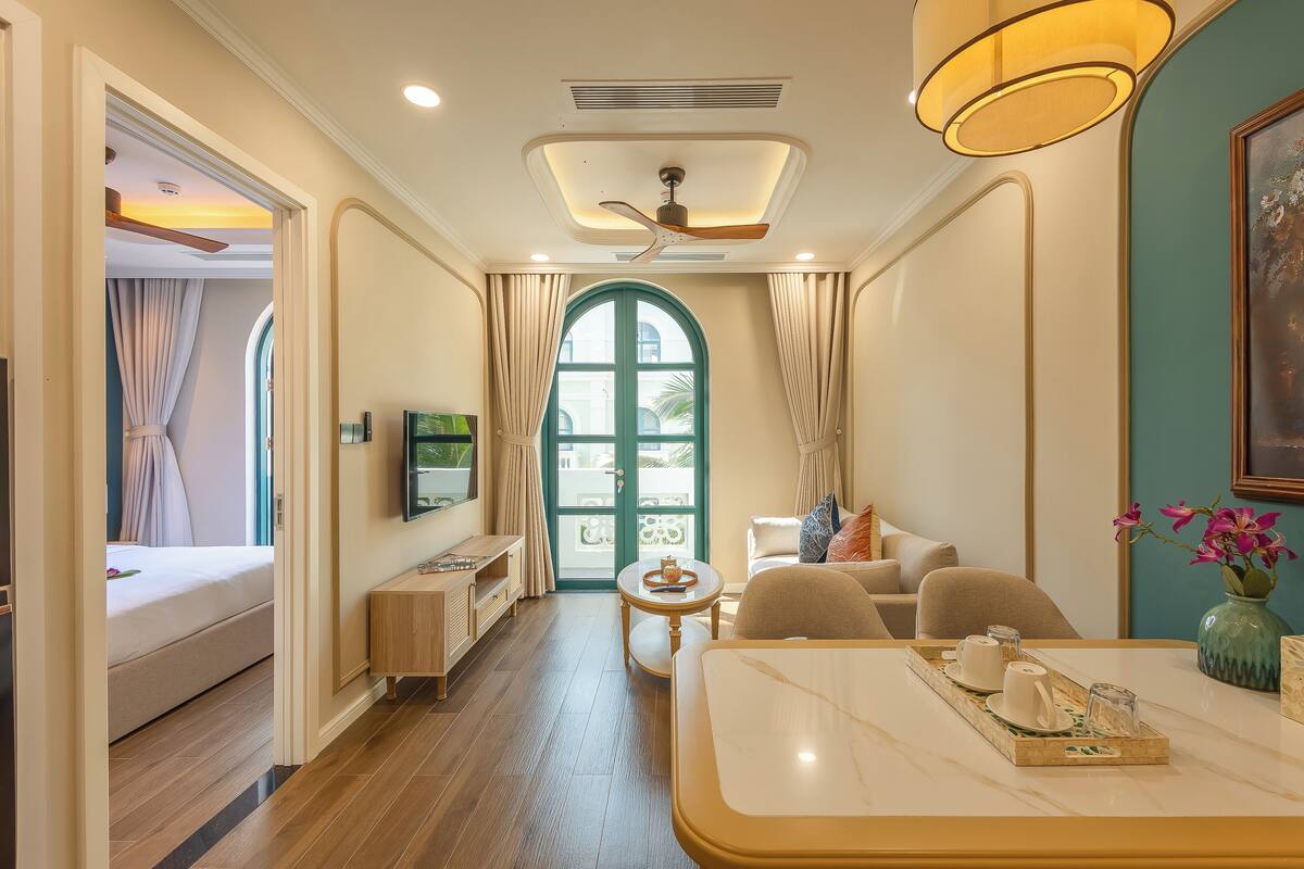 Habitación de hotel con cama, zona de estar con mesa y sillas, televisor y un ventanal grande con cortinas.