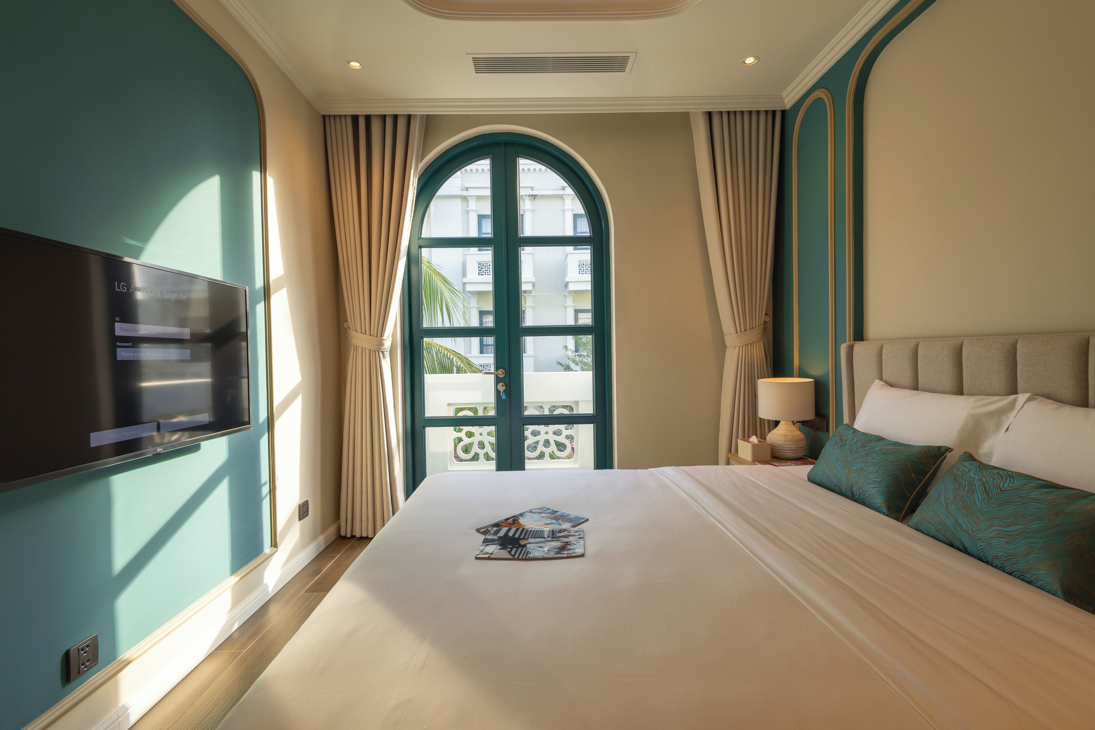 A hotel room with a large bed, a TV on the wall, a window with curtains, and a view of buildings outside.
