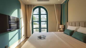A hotel room with a large bed, a TV on the wall, a window with curtains, and a view of buildings outside.