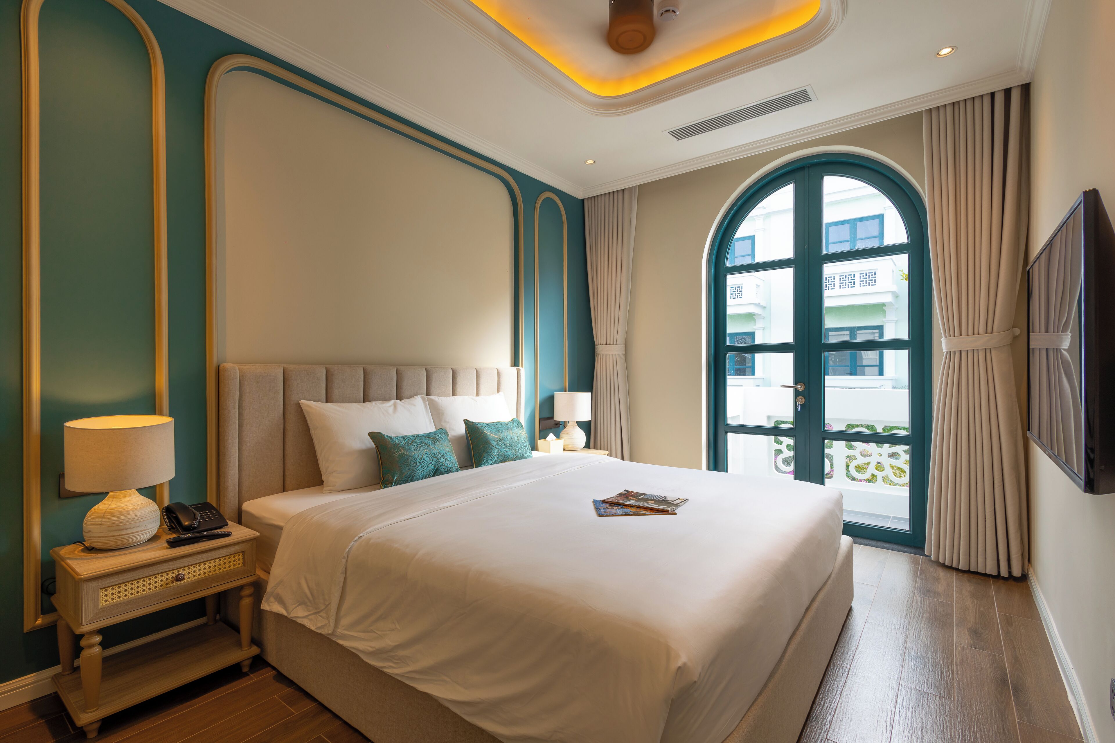 A neatly made bed with a white comforter and pillows, a bedside table with a lamp and phone, and a large window with curtains.