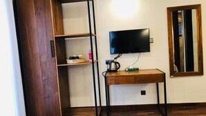 A room with a wooden floor, a TV mounted on the wall, a wooden desk with a chair, and a shelf unit with a mirror. - Hotel Bulwark (Nuwara Eliya)
