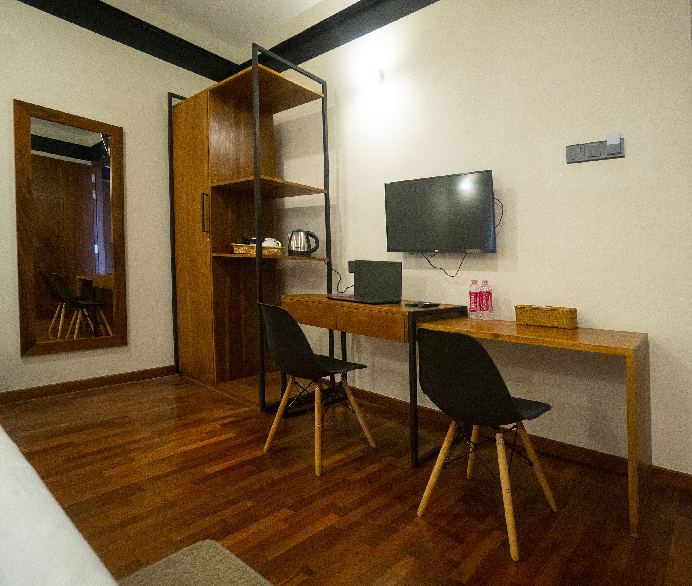 A room with a wooden desk, two black chairs, a television, a shelf with a kettle and a basket, and a wooden wardrobe.