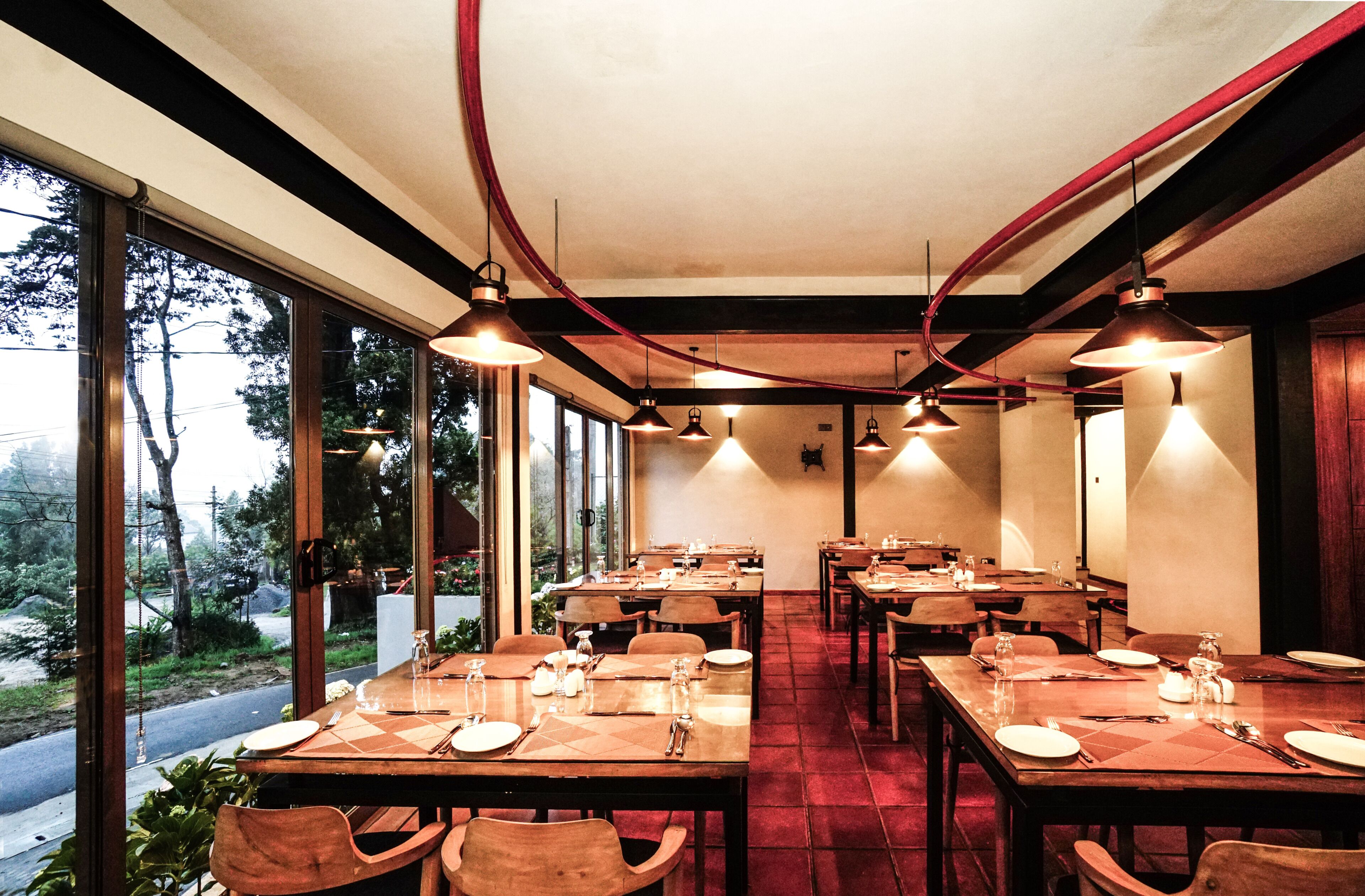 Un restaurante con varias mesas dispuestas para comer, amplias ventanas que ofrecen vistas al exterior y lámparas colgantes.