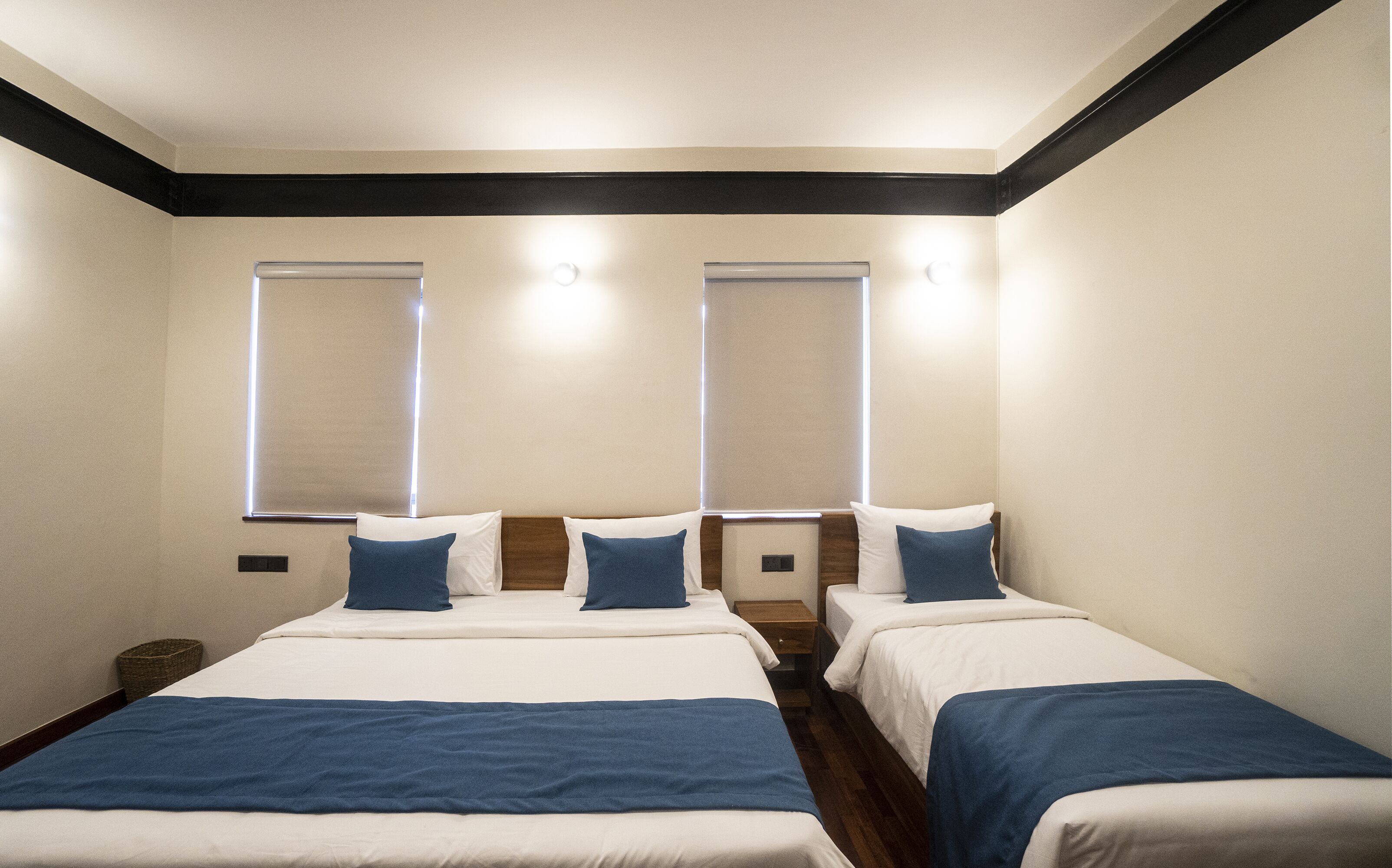 A hotel room with two beds, each with a blue blanket, white bedding, and wooden headboards. There are two bedside tables and a small nightstand. The room has a modern design with a dark trim around the ceiling and walls.