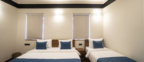 Habitación de hotel con dos camas, cada una con una manta azul, ropa de cama blanca y cabeceros de madera. Hay dos mesitas de noche y una pequeña mesita auxiliar. La habitación tiene un diseño moderno con detalles oscuros en el techo y las paredes.