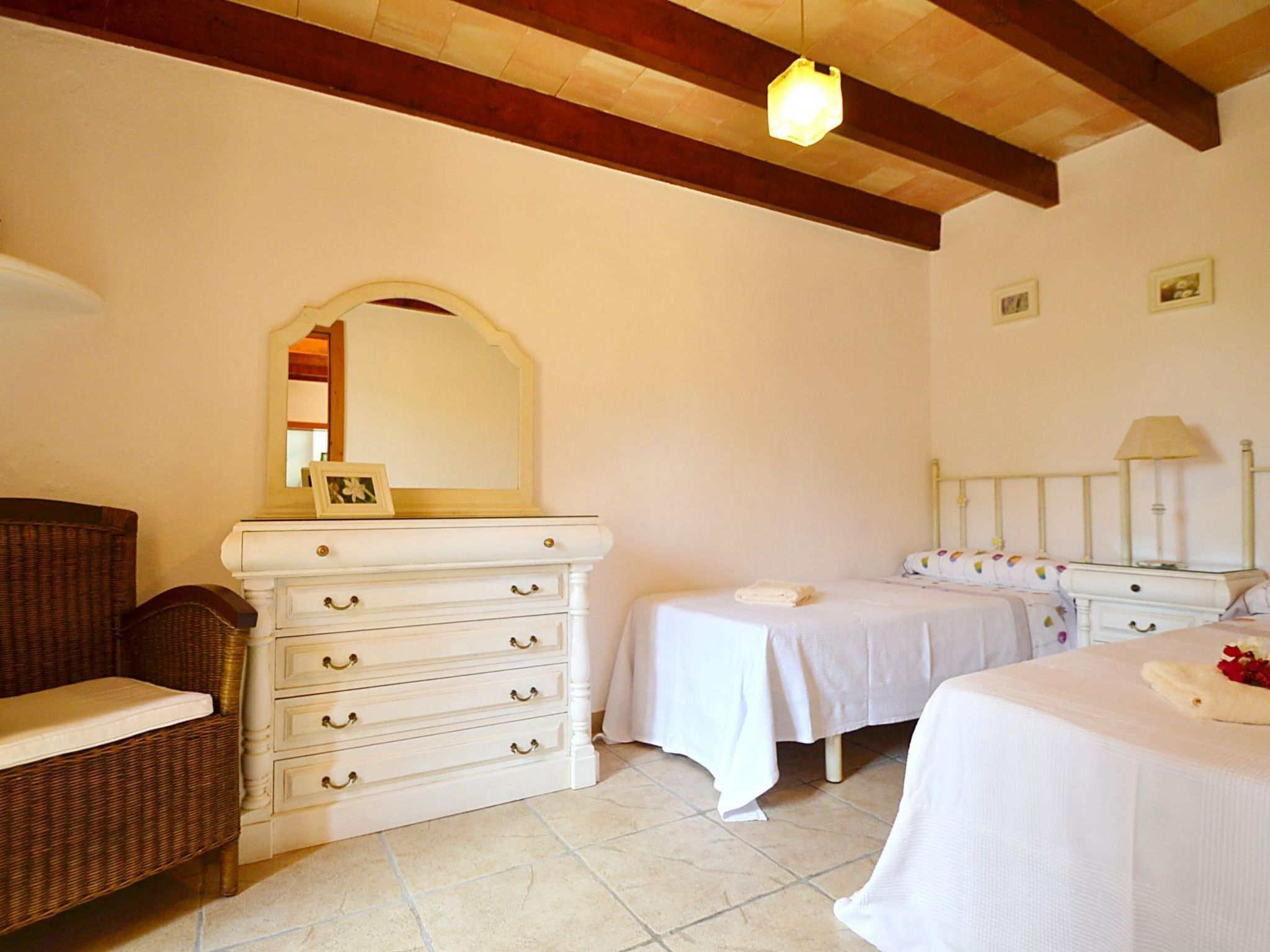 A bedroom with a white dresser, two beds, a wicker chair, and a wooden ceiling.