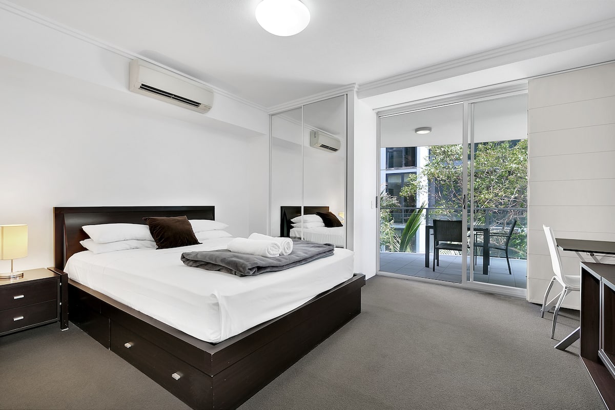 Una habitación de hotel moderna con una cama grande, un escritorio y un balcón con mobiliario exterior.