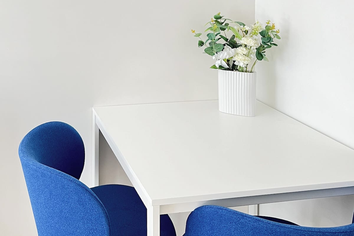 Una mesa de comedor blanca con dos sillas azules, un jarrón blanco con flores blancas y un espejo redondo con marco azul en la pared.