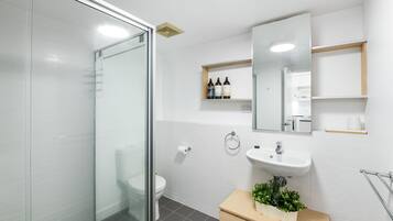 A modern bathroom with a glass shower enclosure, a white toilet, a sink with a mirror above, and a small shelf with toiletries.
