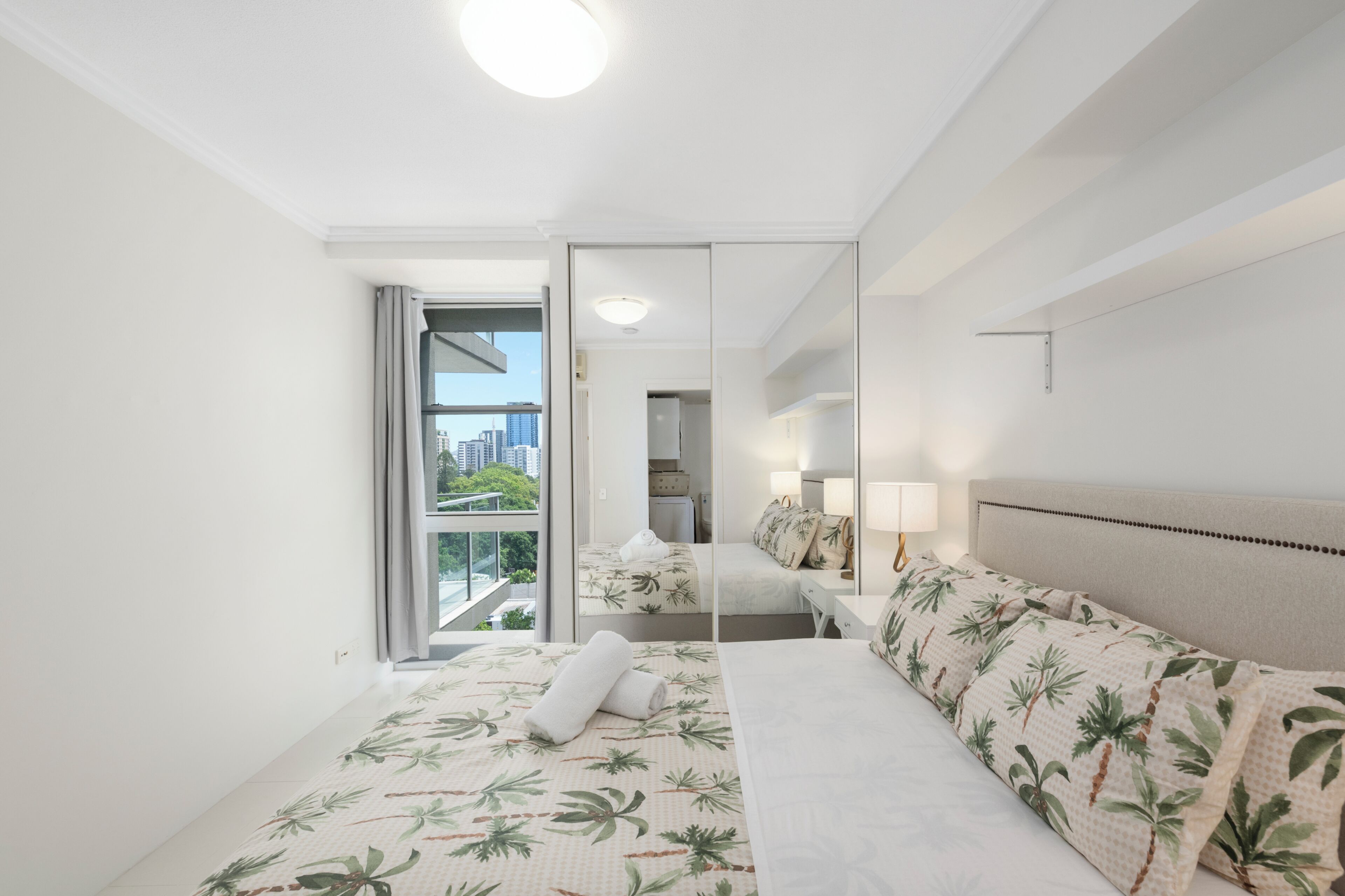A hotel room with a bed, a nightstand, a lamp, a mirror, and a view of the city.