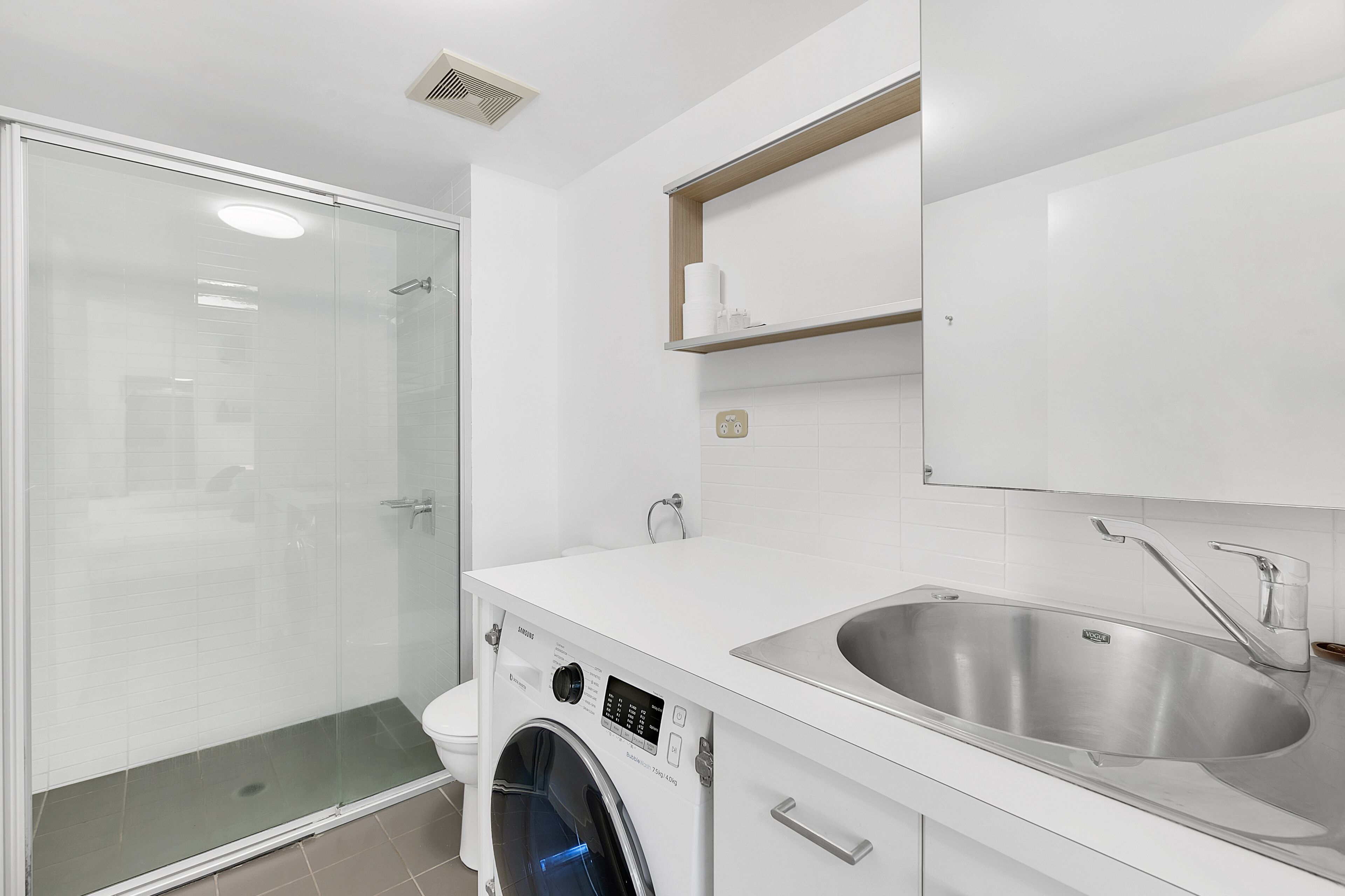 A modern bathroom with a glass-enclosed shower, a built-in washing machine, and a stainless steel sink.