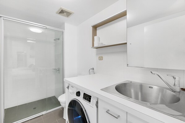 Un baño moderno con ducha de vidrio, lavadora integrada y lavamanos de acero inoxidable.