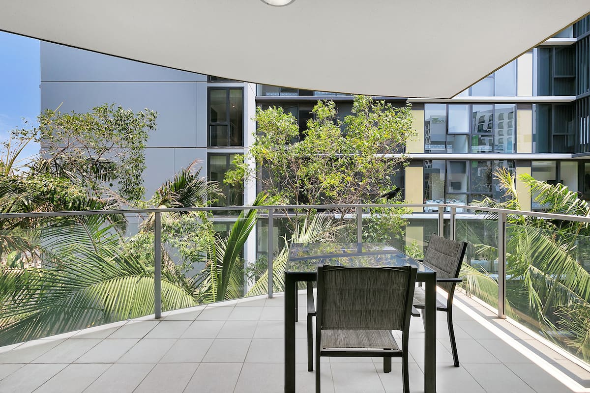 Un espacio exterior moderno con mobiliario de jardín, una mesa y sillas de vidrio, rodeado por una barandilla de cristal, con vista a un edificio con grandes ventanales y plantas.