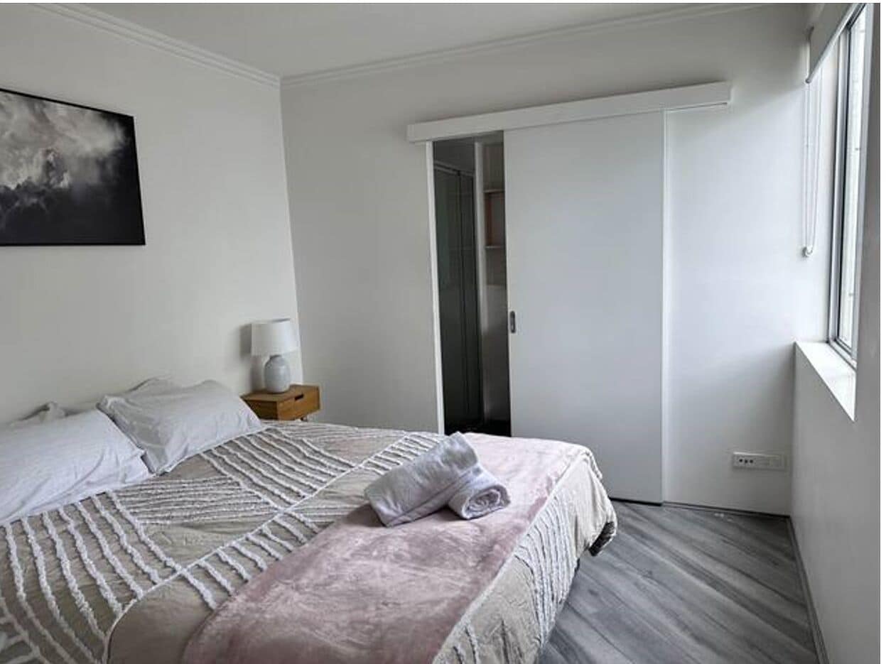 Habitación de hotel con una cama, una mesita de noche, una lámpara, un cuadro en la pared y una puerta que da a otra habitación.