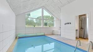 A modern indoor swimming pool with a clear view of the outdoors through large windows, a built-in bench, and a separate shower area.
