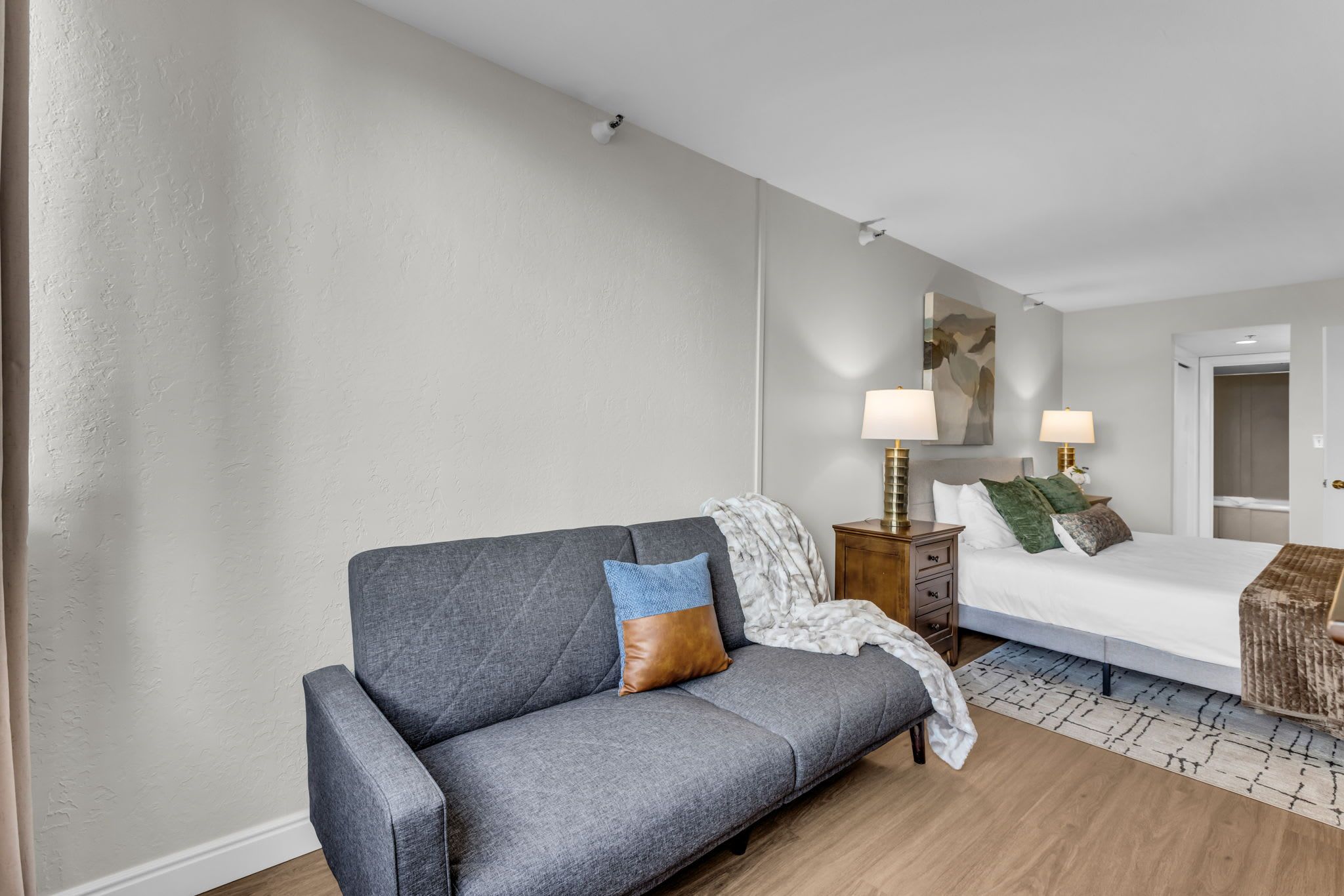 Un dormitorio moderno con un sofá gris, una mesita de noche de madera, una cama con ropa de cama blanca y un espejo grande en la pared.