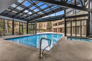 Piscina interior con techos de vidrio transparente, un área de descanso con sillones y vistas a edificios exteriores.