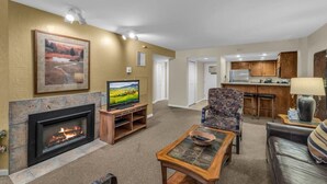 A living room with a fireplace, a television, a patterned armchair, a sofa, and a kitchen area in the background.