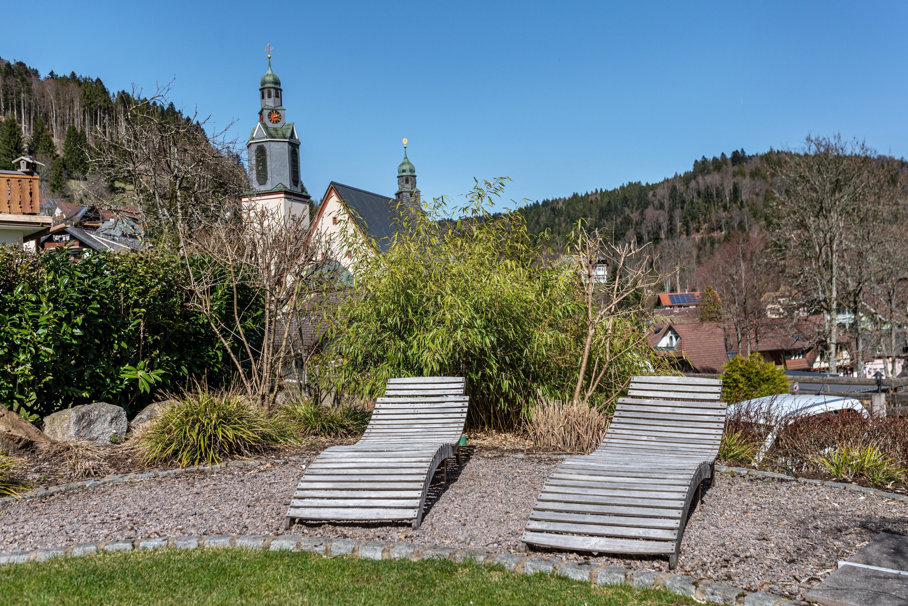 A serene outdoor area with two wooden loungers, a church with a clock tower, and a mountainous backdrop.