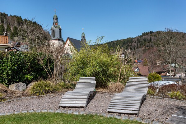 A serene outdoor area with two wooden loungers, a church with a clock tower, and a mountainous backdrop.