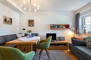 A modern living room with a wooden dining table, green chairs, a sofa, a TV, bookshelves, and a window with curtains.