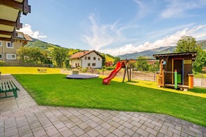 A playground with a slide, a trampoline, and a small wooden structure, set against a backdrop of a house and mountains.