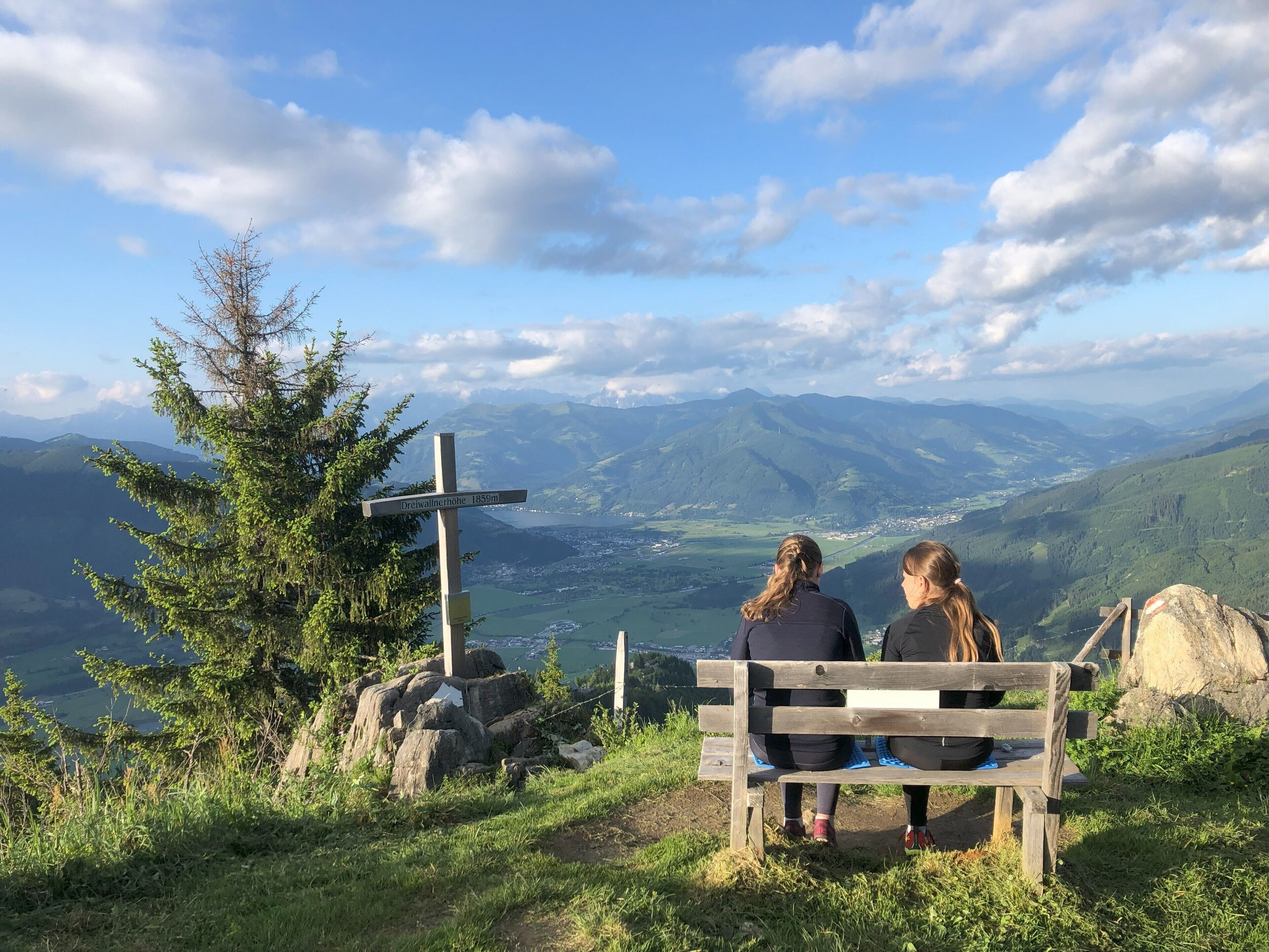 Zwei Personen sitzen auf einer Holzbank und blicken auf eine gebirgige Landschaft. Ein Wegweiser zeigt auf einen Aussichtspunkt.