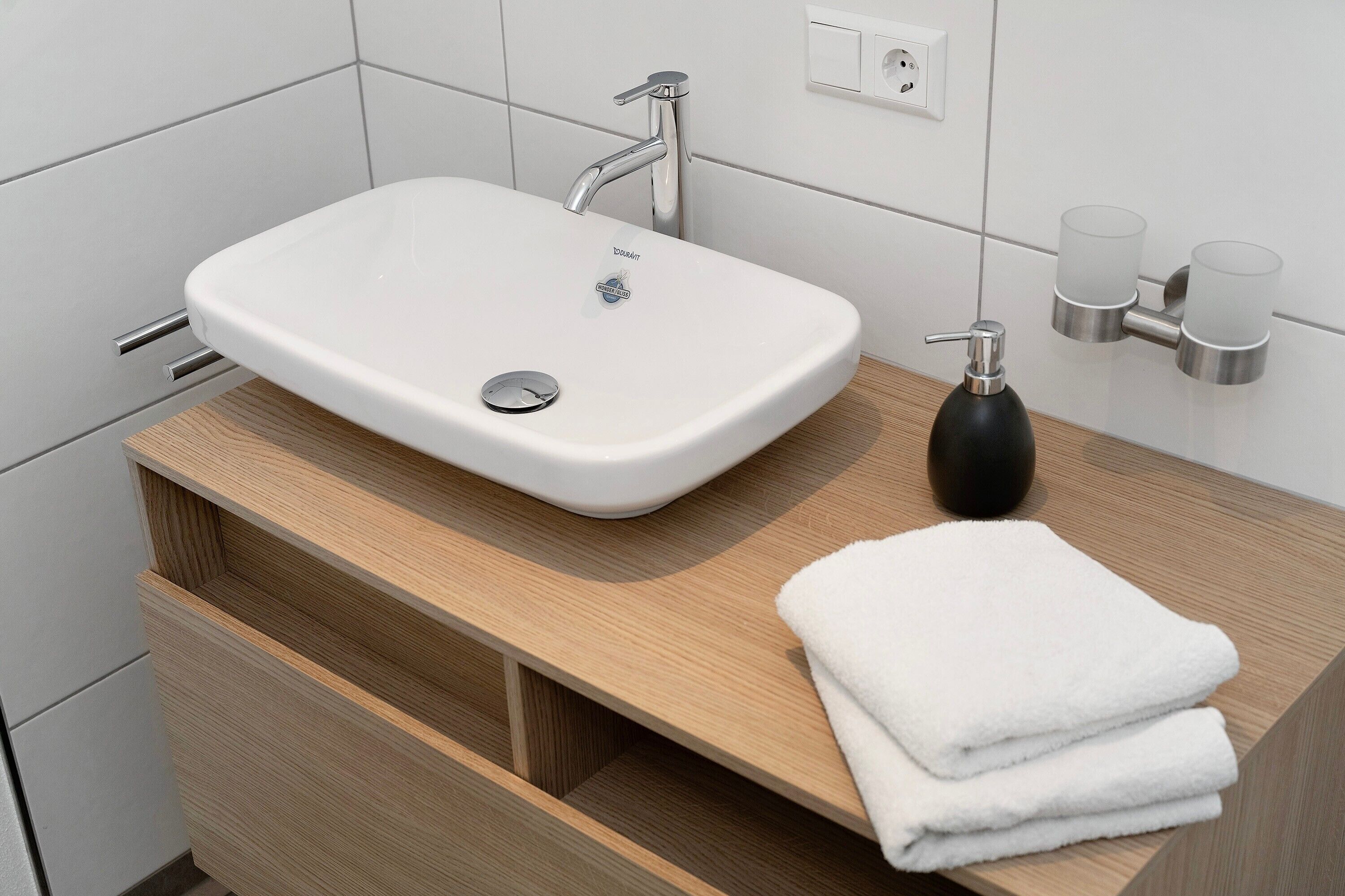 Ein modernes Badezimmer mit einem weißen Waschbecken, einem Holzschranken und gestapelten Handtüchern.