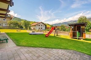 Un terrain de jeux avec un toboggan, un trampoline et une petite structure en bois, le tout situé devant une maison et des montagnes.