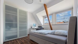 A bedroom with a bed, a desk, a computer, and a window with a view of the sky.