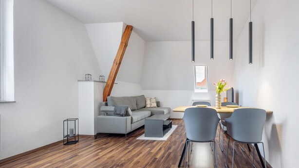 A modern living room with a wooden floor, grey sofa, dining table with chairs, pendant lights, and a sloped ceiling.