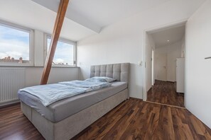 A bedroom with a bed, wooden flooring, a window, and a door leading to another room.