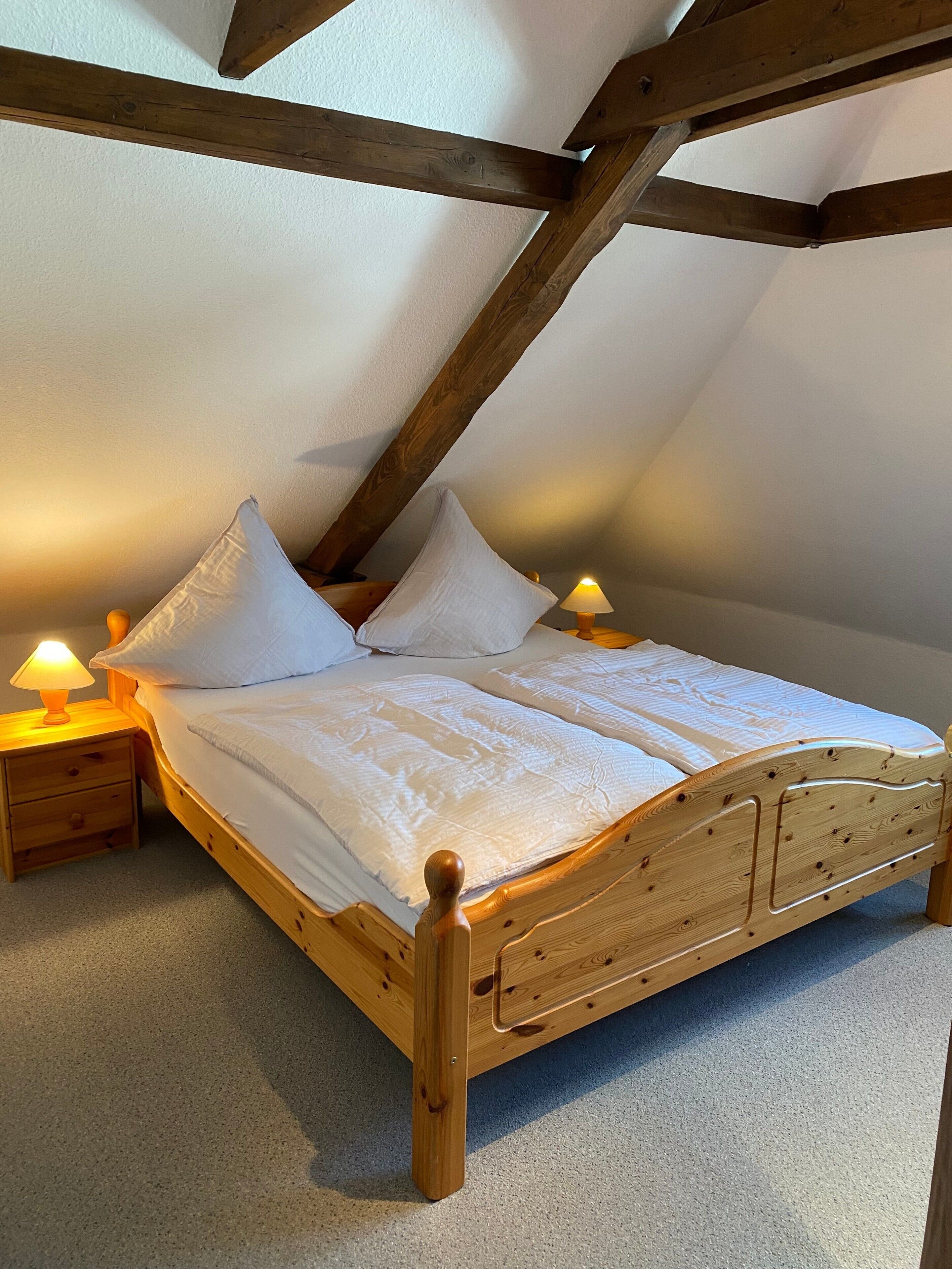 A wooden bed with a headboard, two bedside tables with lamps, and a sloped ceiling with wooden beams.