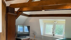 A loft-style room with exposed wooden beams, a television, a sewing machine, a green sofa, and a window with a view of greenery.