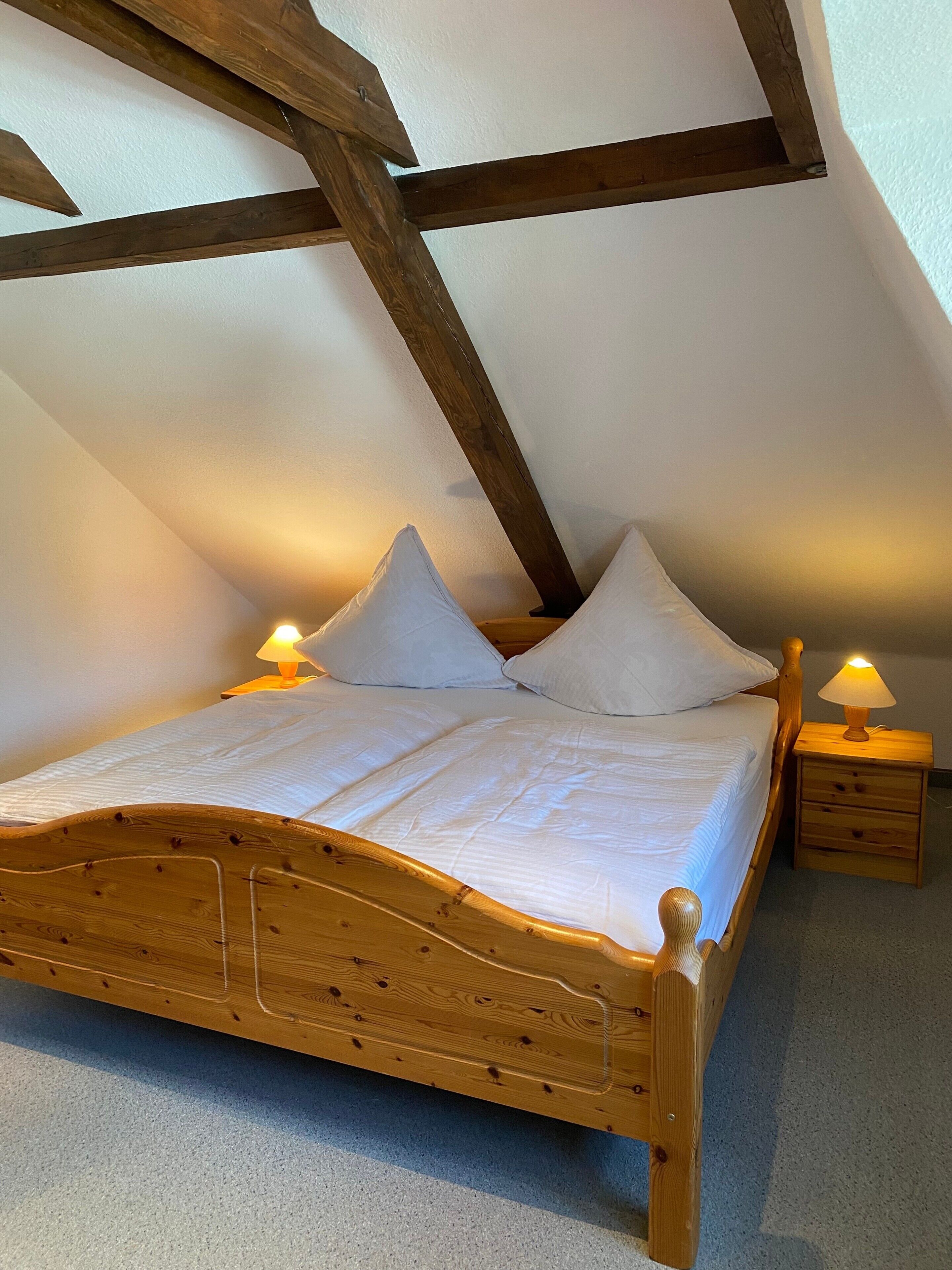 Een houten bed met wit beddengoed in een kamer met schuine plafonds en zichtbare balken.
