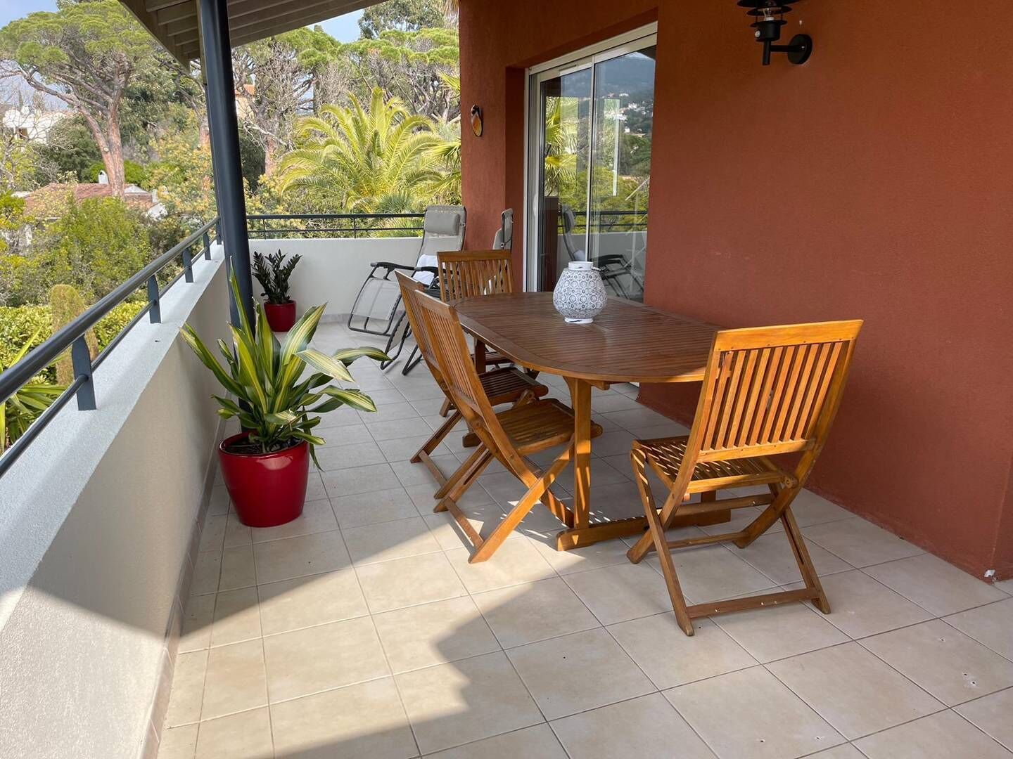 Uma varanda com mesa e cadeiras de madeira, plantas em vasos e vista para árvores e edifícios.